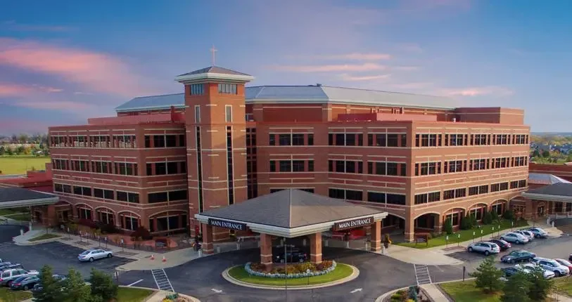 Large red brick building with multiple floors, main entrance marked, surrounded by parking lots and green spaces, under a partly cloudy sky.