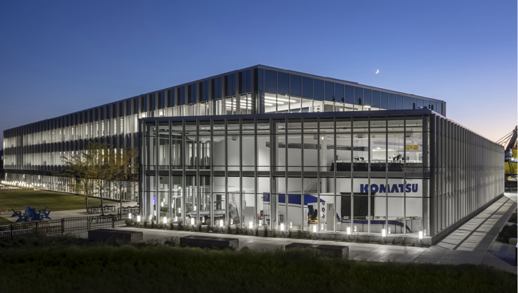 Modern glass building with aircraft inside and company logo 'KONAMTSU,' illuminated at dusk.