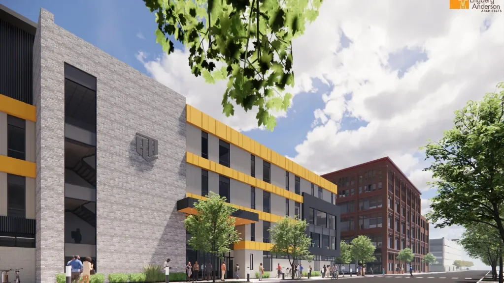 A modern multi-story commercial building with yellow accents, large windows, and a brick exterior, surrounded by trees and people walking on the sidewalk.