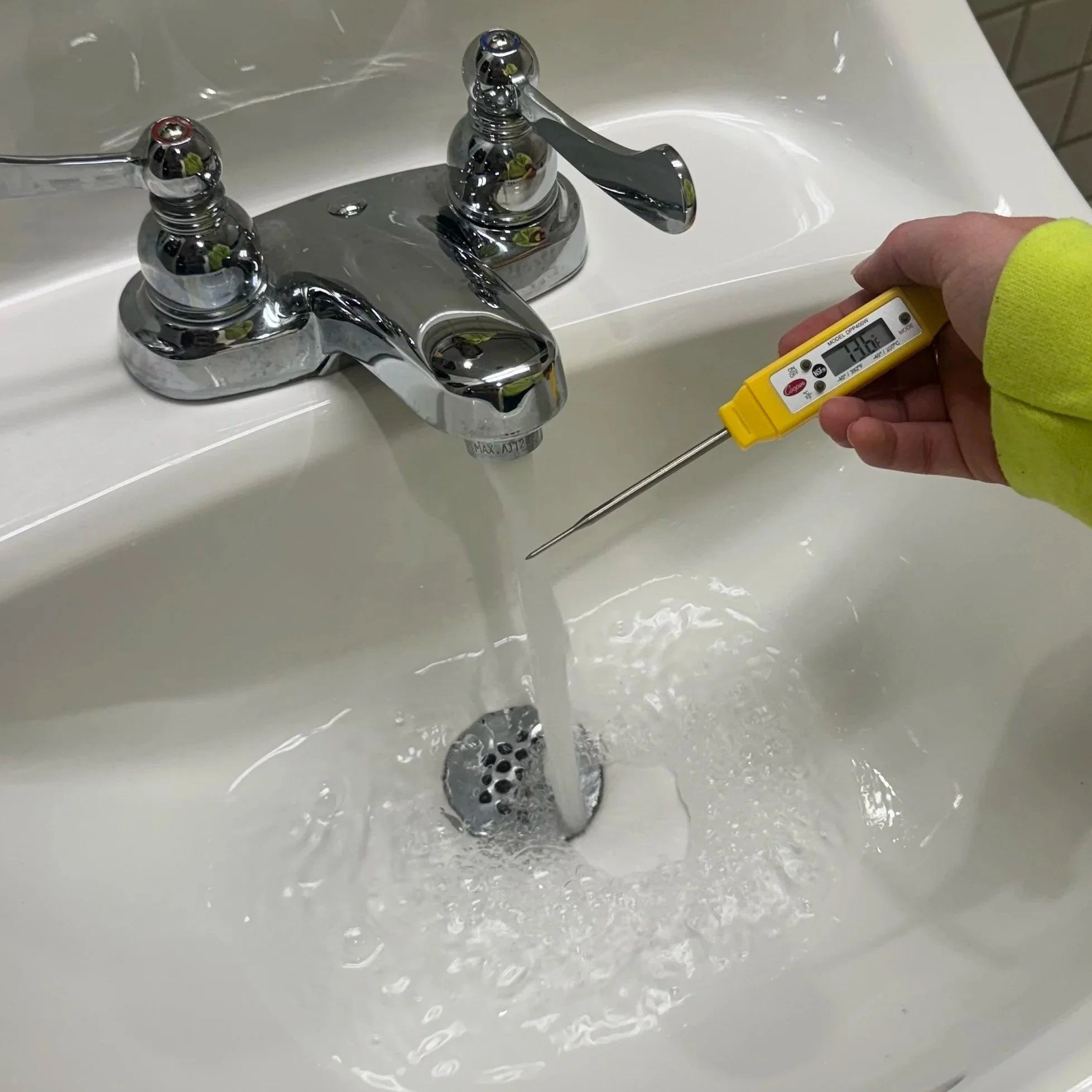 BTI Balance Technologies worker holding a digital thermometer showing a temperature of 73.6°F near a bathroom sink with a chrome faucet and running water to ensure correct temperatures in domestic waters. .