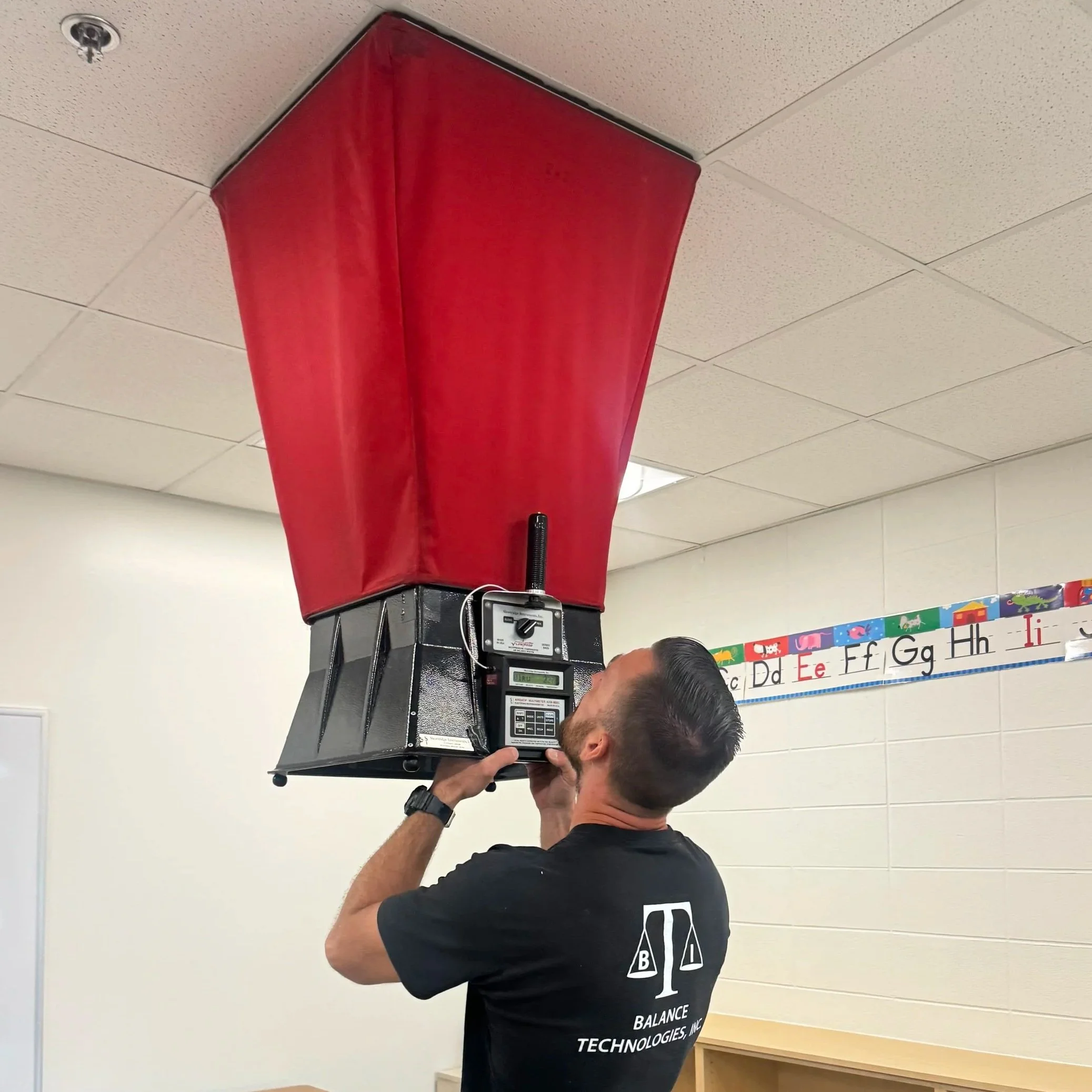 Balance Technologies (BTI) worker holding air hood apparatus taking air flow measurements and balancing in a classroom.