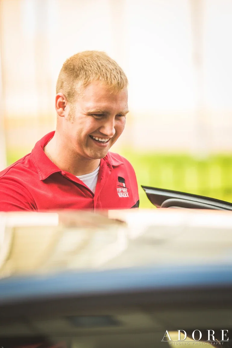A valet, smiling, wearing a red uniform with a logo, at a cashier counter in a retail store.