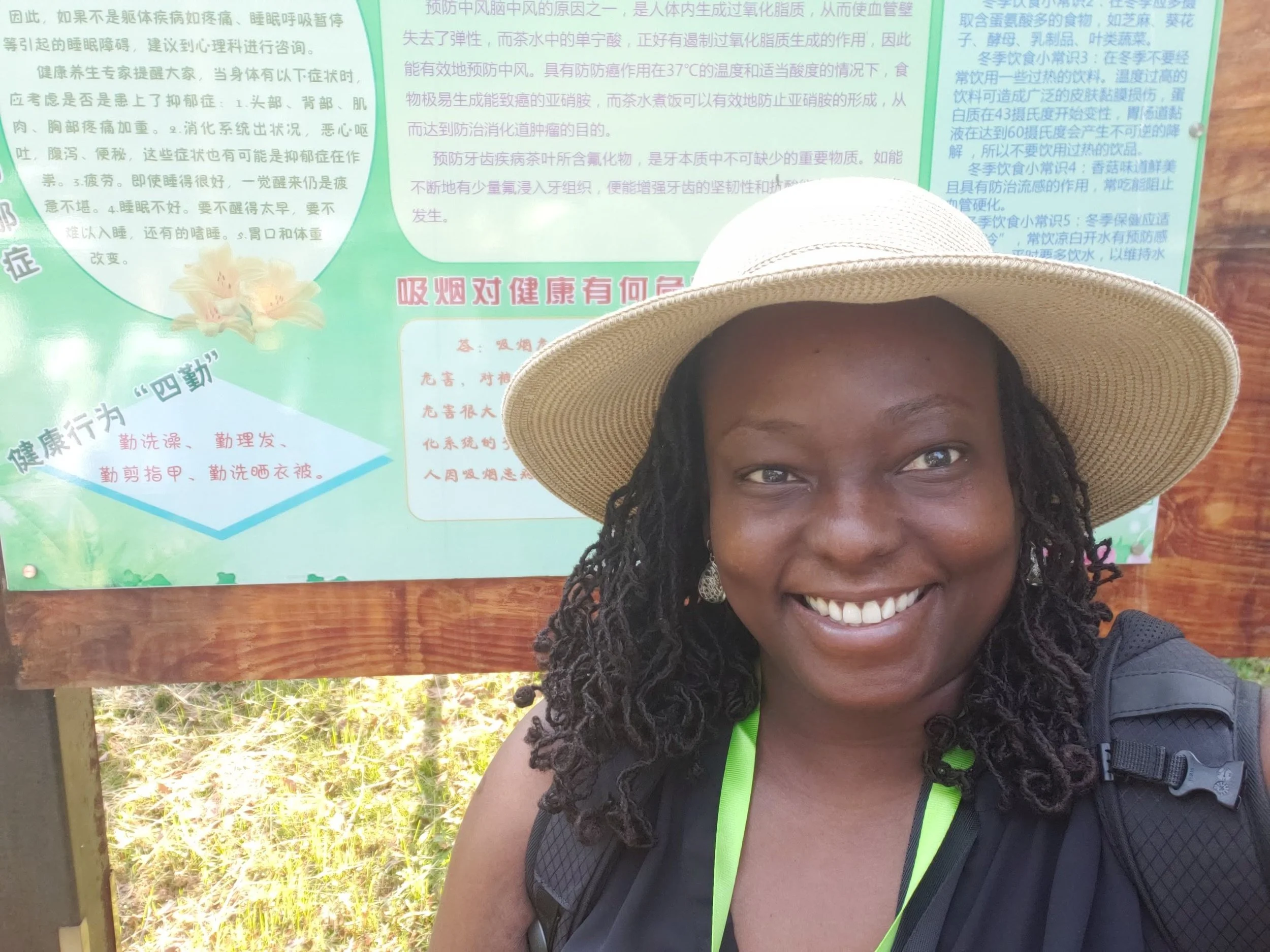 Close-up of a smiling woman with dark braided hair wearing a wide-brimmed hat, with a colorful informational poster in Chinese in the background. The woman is outdoors, sitting at a wooden table, and the photo captures her face from the nose up.