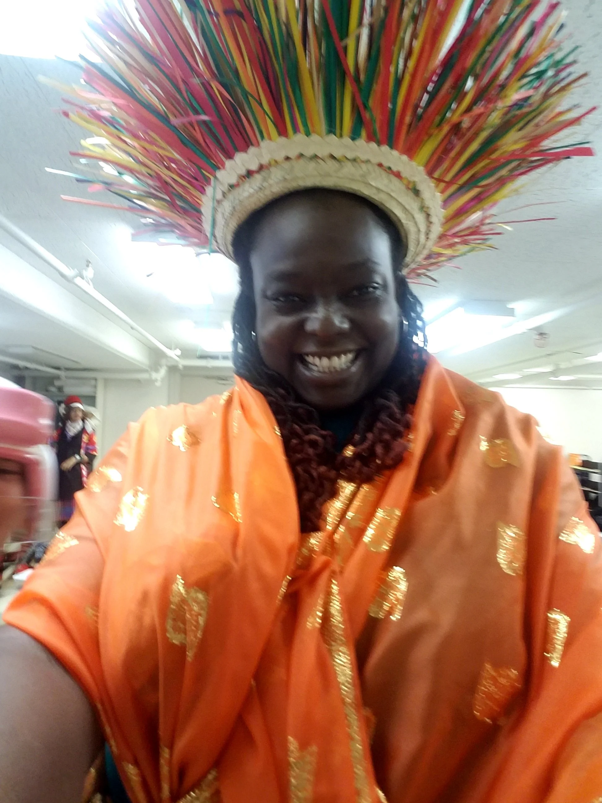 A woman smiling while wearing a colorful traditional headdress and an orange outfit with gold accents, indoors with a person in the background.