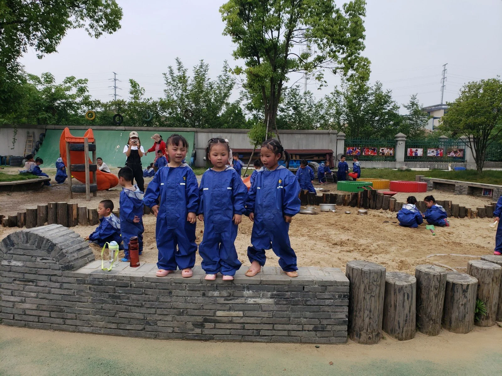 Young children in blue uniforms playing and sitting on a playground with trees and a fence in the background.