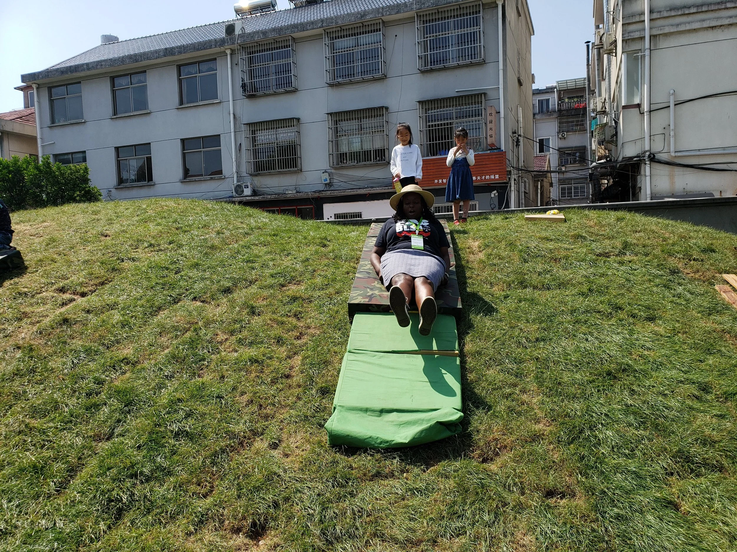 Three children on a grassy hill, two girls standing and a girl lying on a padded mat. Buildings with barred windows are in the background.