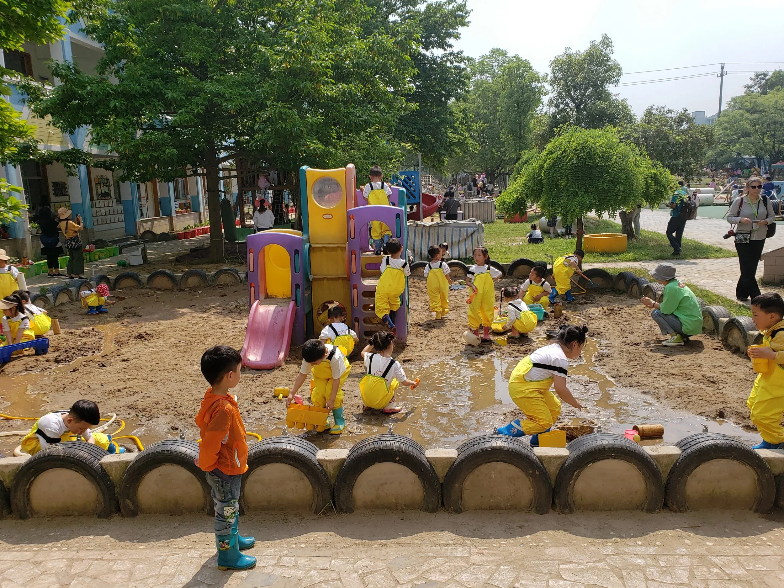Children playing in a sandbox area at a playground, surrounded by tire barriers, with trees and other playground equipment in the background.