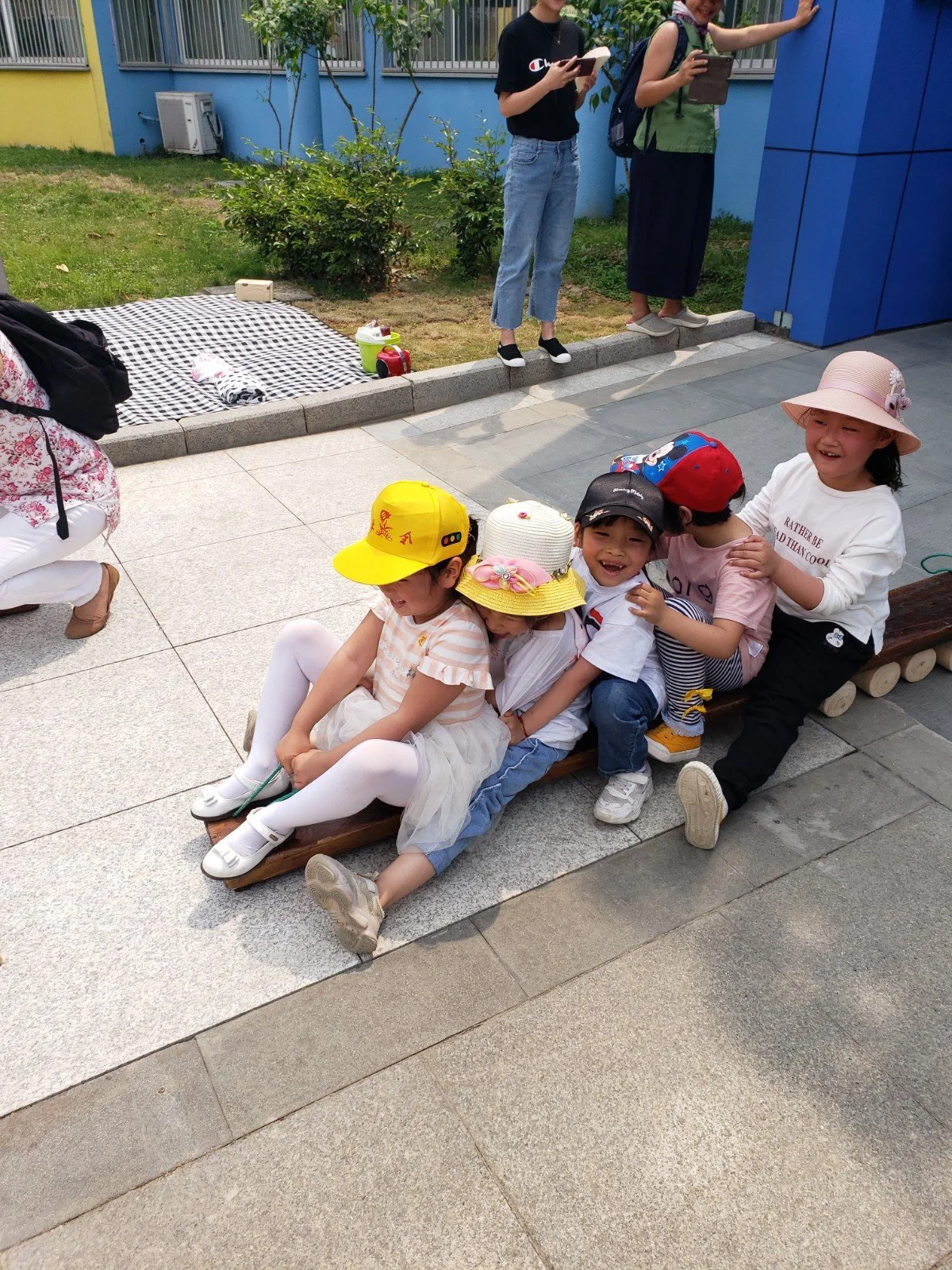 A group of young children sitting on a wooden bench, smiling and embracing each other, wearing hats and casual clothes in an outdoor area with a paved walkway and a grassy background.