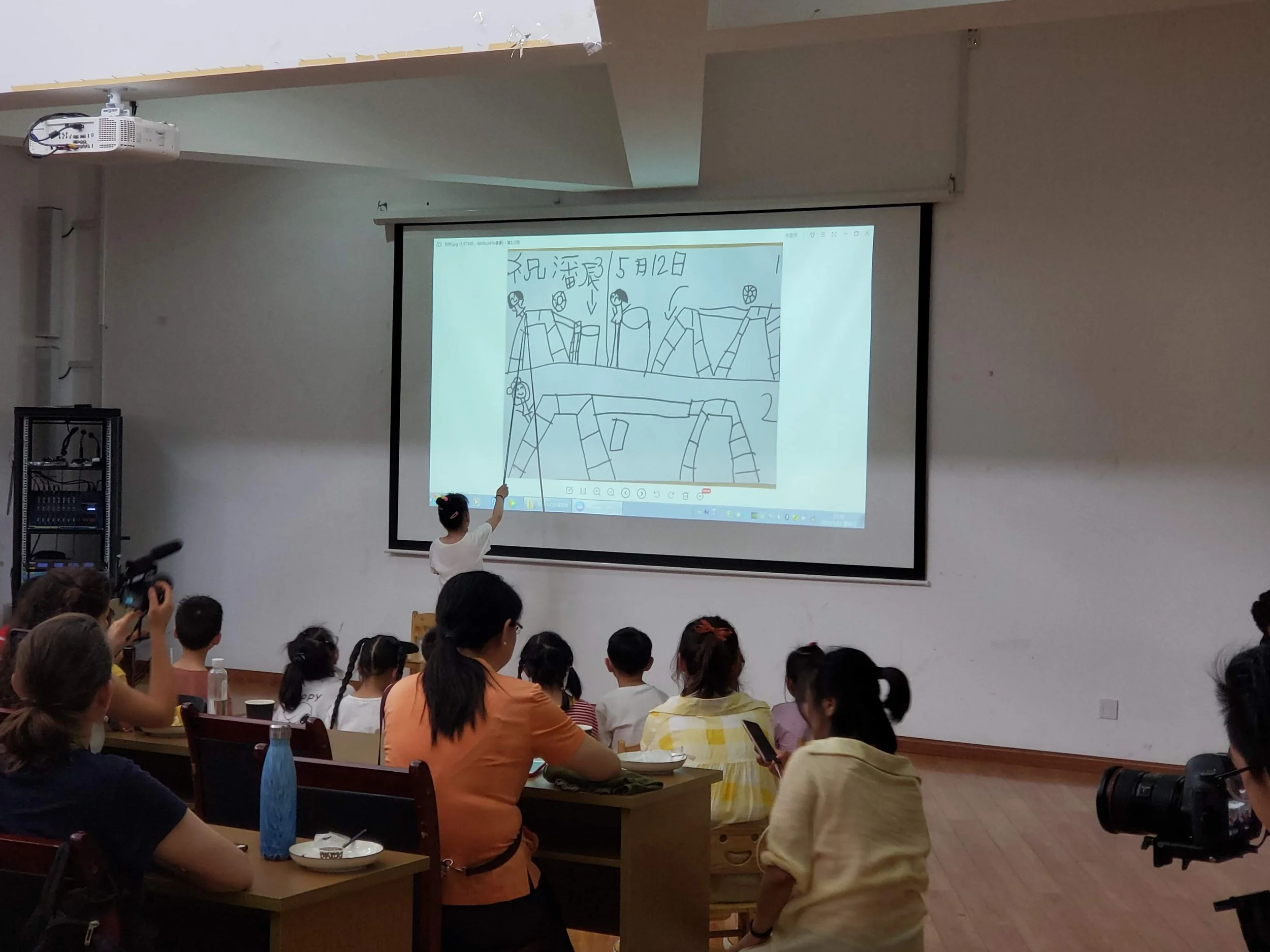 A classroom or lecture hall filled with people watching a presentation on a large screen. The slide on the screen shows a child's drawing of people and some Japanese text. A child is pointing at the screen, and there is a projector mounted on the ceiling.