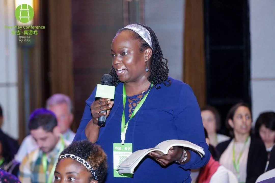 A woman wearing a blue dress speaking into a microphone at a conference, holding a book, with attendees in the background and a green conference badge around her neck.