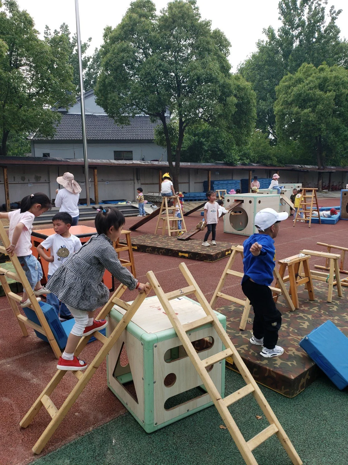 Children playing outdoors on a playground with climbing structures, slides, and other play equipment, under trees on a cloudy day.