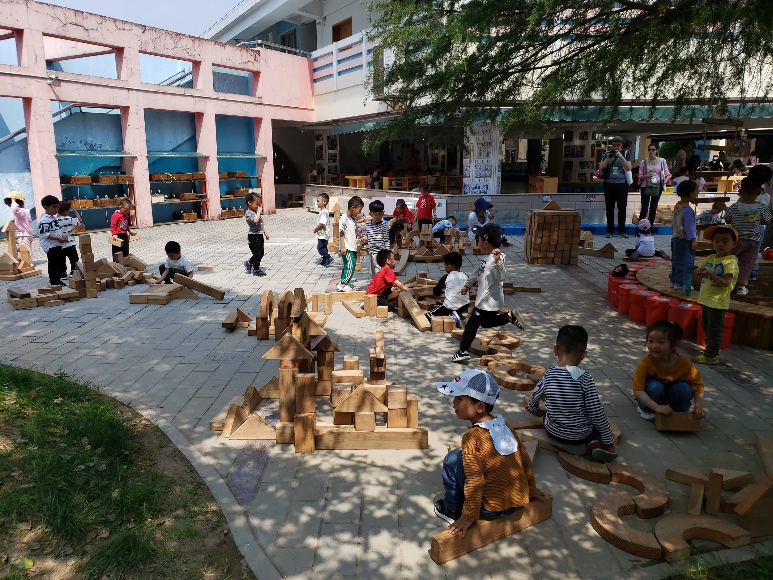 Children playing with large wooden building blocks outdoors at a courtyard area near a building with pink and white walls and a shaded tree overhead.