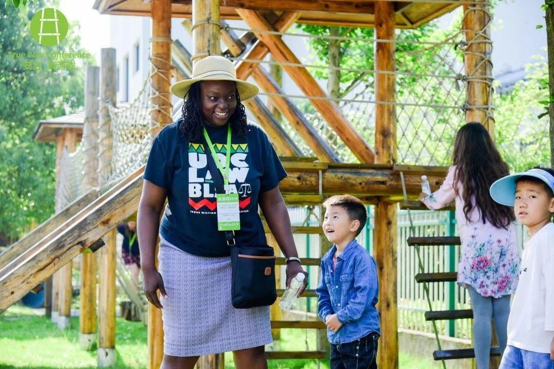 A woman with a hat smiling and talking to a young boy in a blue jacket at an outdoor playground with wooden structures, surrounded by greenery and children playing.