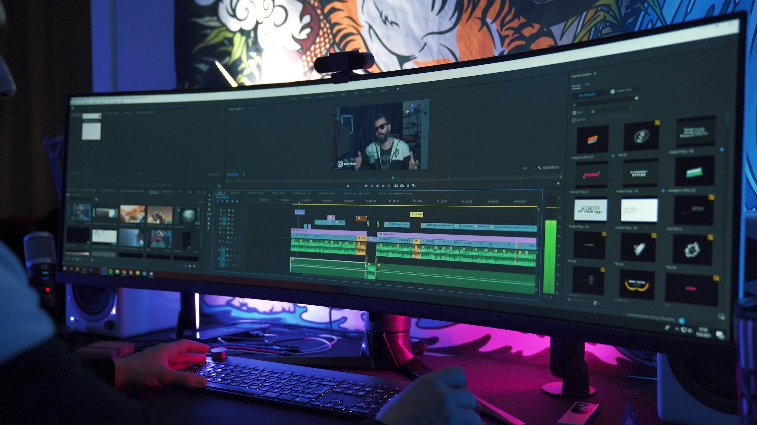 A person working on video editing at a computer with a large curved monitor displaying a timeline in editing software. The workspace has colorful lighting and artwork on the wall in the background.