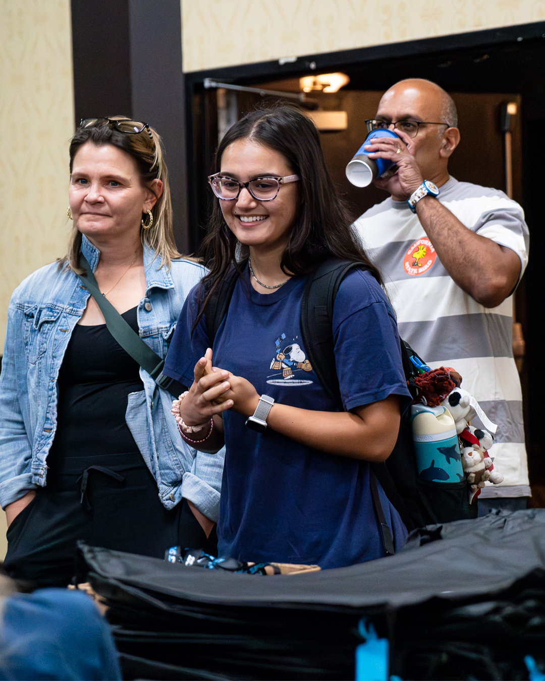 Group of three people standing indoors, smiling, with a young woman at the center wearing glasses and a backpack, and two adults on either side, one drinking from a blue cup.