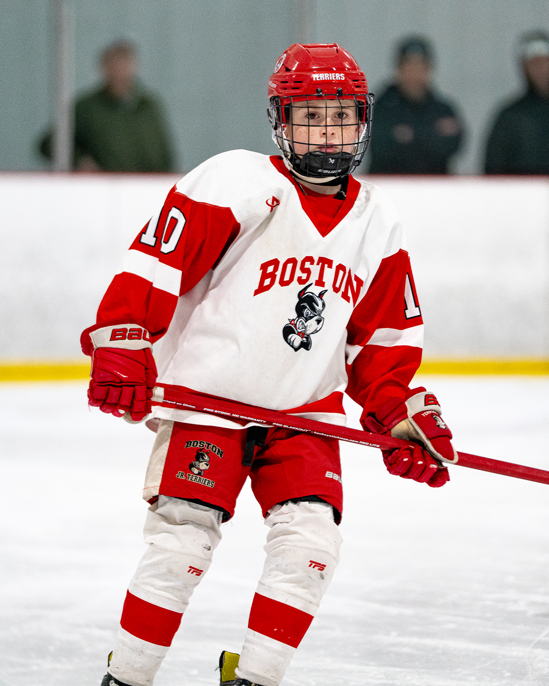 Young male hockey player wearing a white and red Boston Jr. Terriers uniform, red helmet, and holding a red hockey stick on the ice rink.