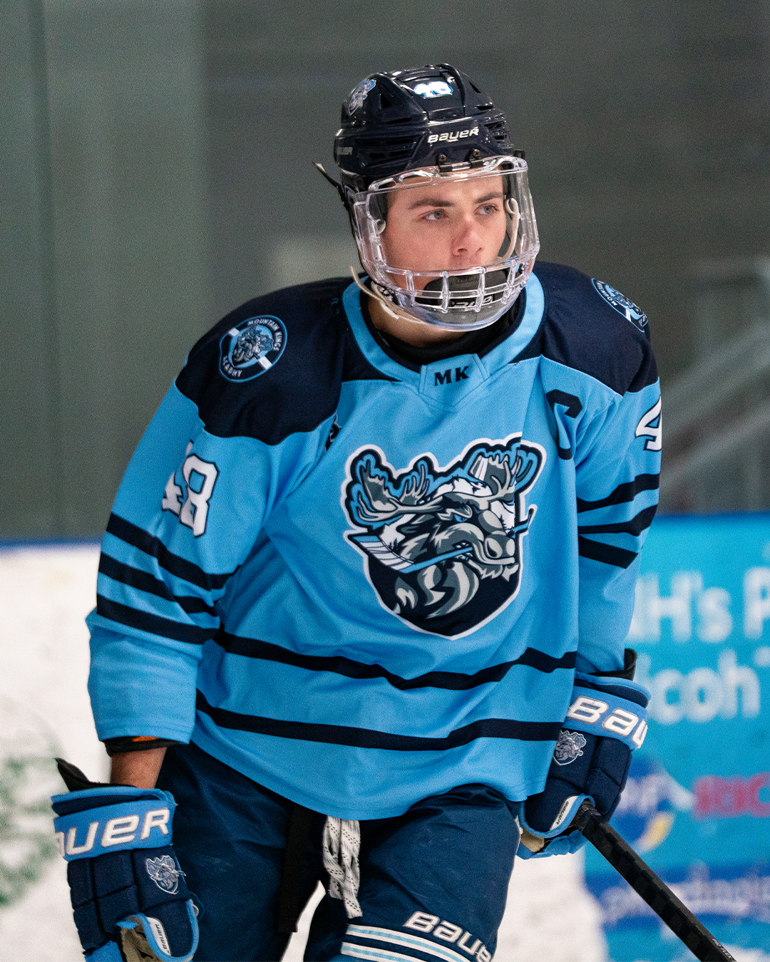 Young male ice hockey player in a blue jersey with a moose logo, wearing a helmet and gloves, standing on the ice rink during a game.
