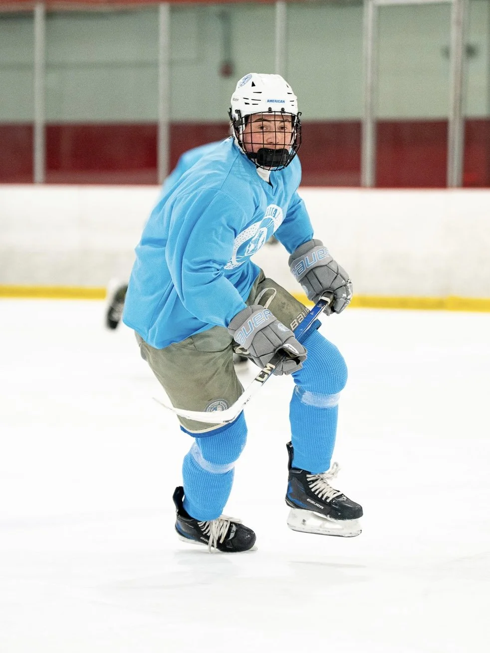 A hockey player on ice rink, wearing a blue jersey, helmet, gloves, and skates, holding a hockey stick, with a playful facial expression.