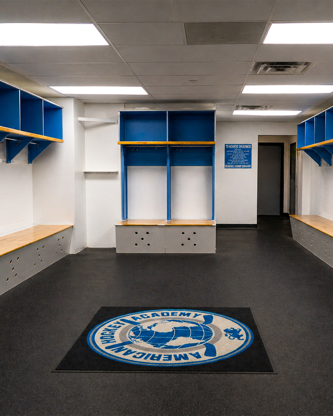 Empty locker room with blue and wood benches, white and blue walls, and a gym logo mat reading 'American Hockey Academy' on the black carpeted floor.