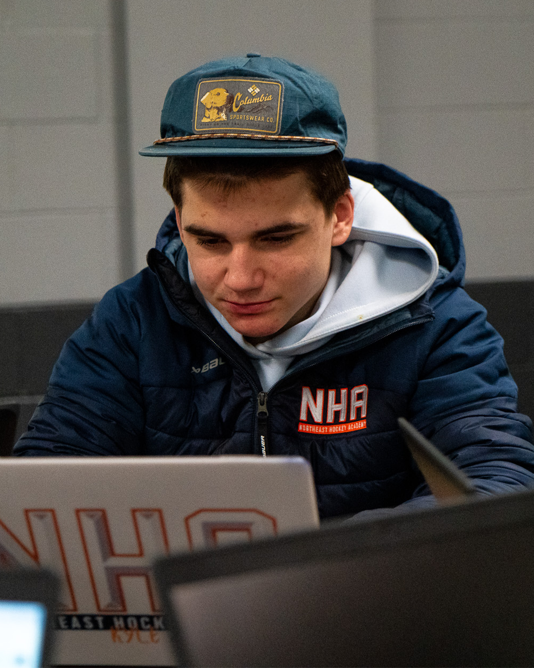 A young man wearing a blue cap with a Columbia Sportswear logo, a navy jacket with the NHL logo, and a white hoodie, sitting in front of a laptop, looking down at the screen.