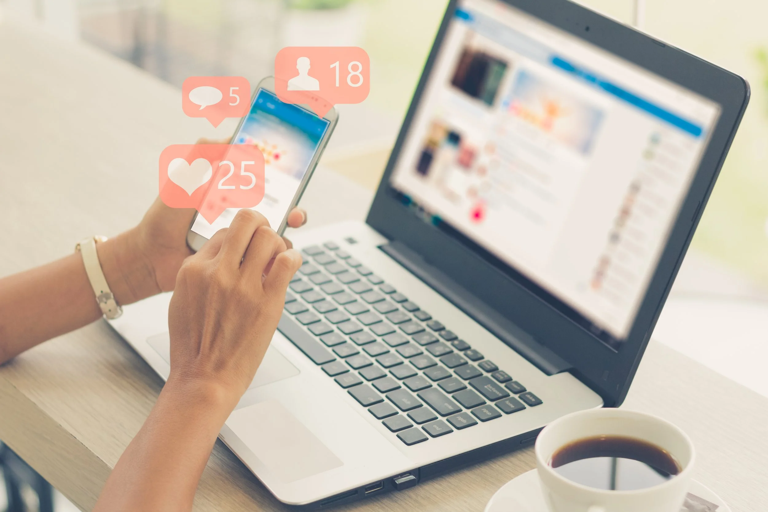 Person holding a smartphone with social media notification icons for messages, friend requests, and likes on its screen, in front of a laptop with social media feed, during daytime.