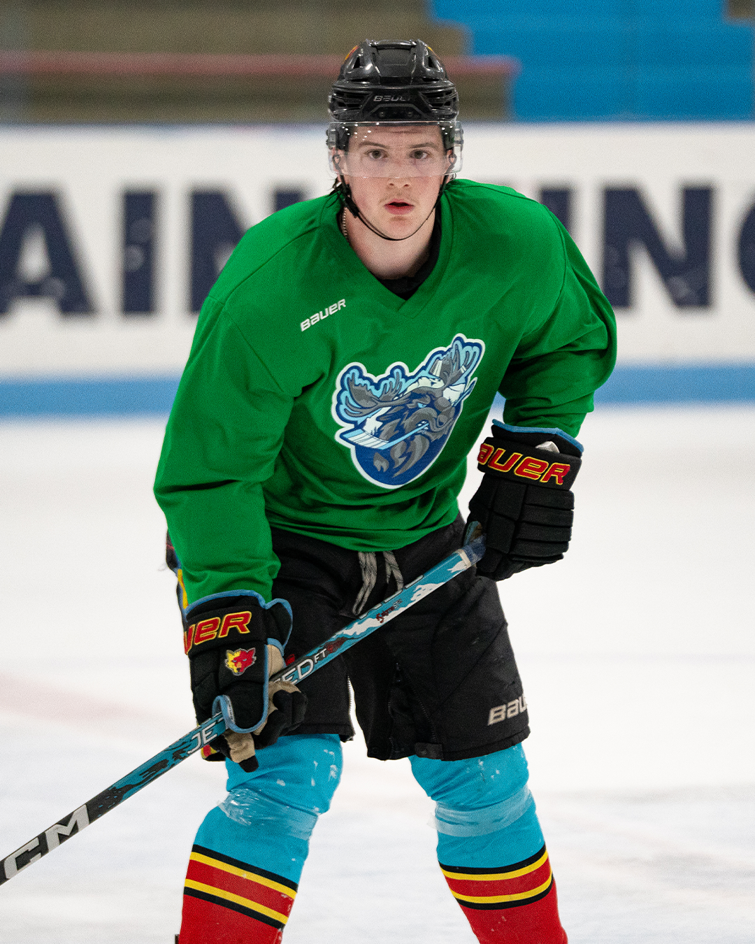A young male ice hockey player in a black helmet, green jersey with a mountain kings logo, black shorts, and blue and red socks, holding a hockey stick on the ice rink.