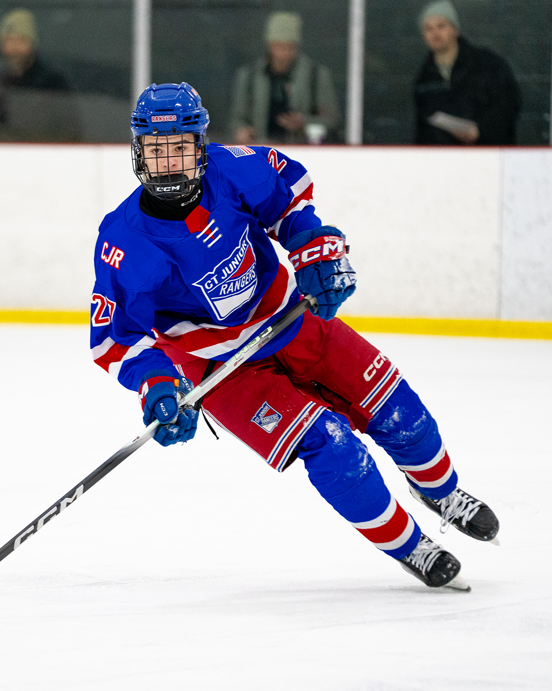 Hockey player in blue and red Gret Rangers jersey, number 27, skating on ice rink with spectators in background.