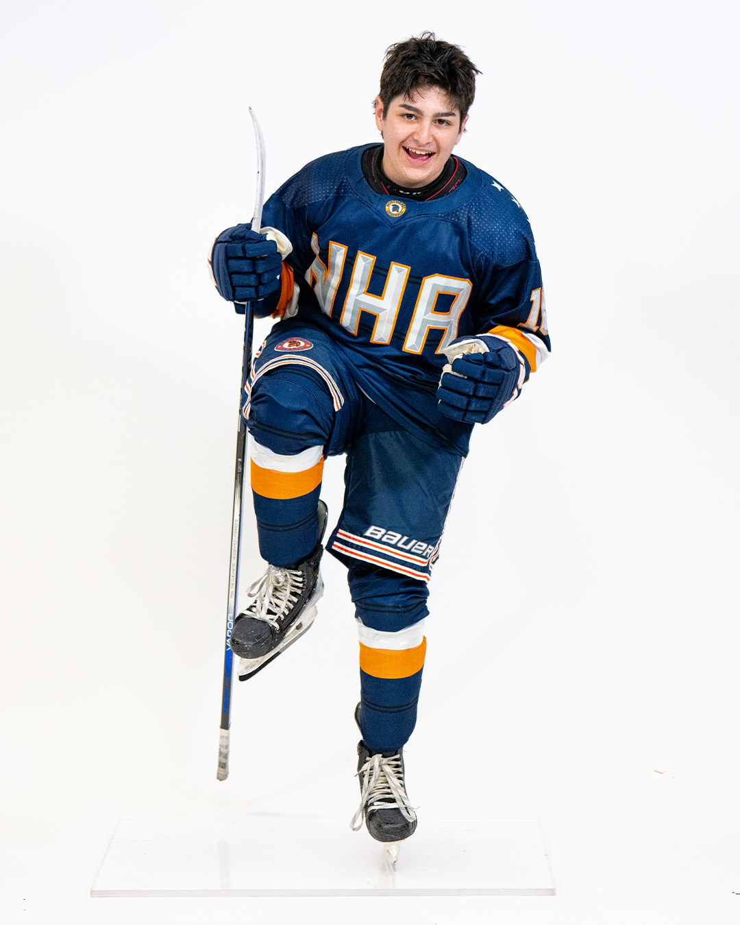 Young male ice hockey player in a dark blue uniform with orange and white accents, posing with one leg raised and a smile, holding a hockey stick against a plain white background.
