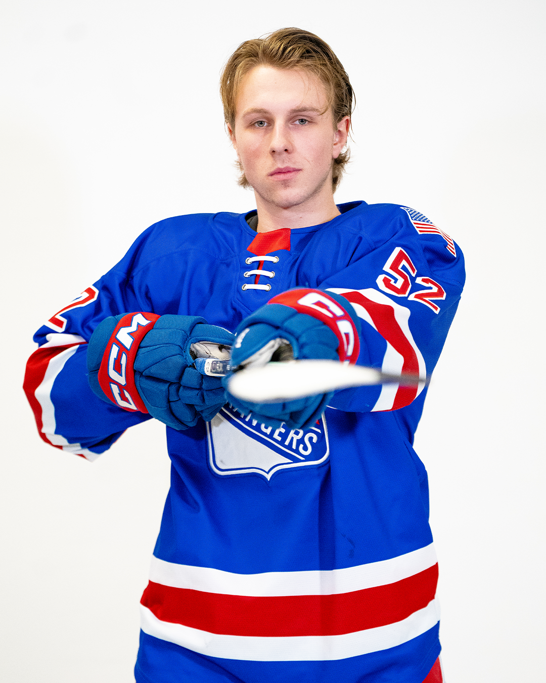 A young male hockey player with blonde hair in a blue University of New York Rangers jersey, holding a hockey stick and wearing blue gloves, standing against a white background.