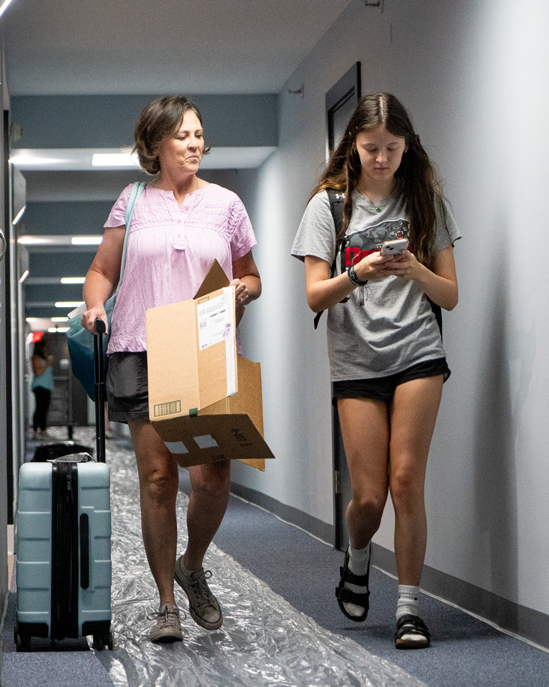 A woman with a rolling suitcase and a girl walking down a hallway, the girl is looking at her phone, wearing casual clothes and sandals.