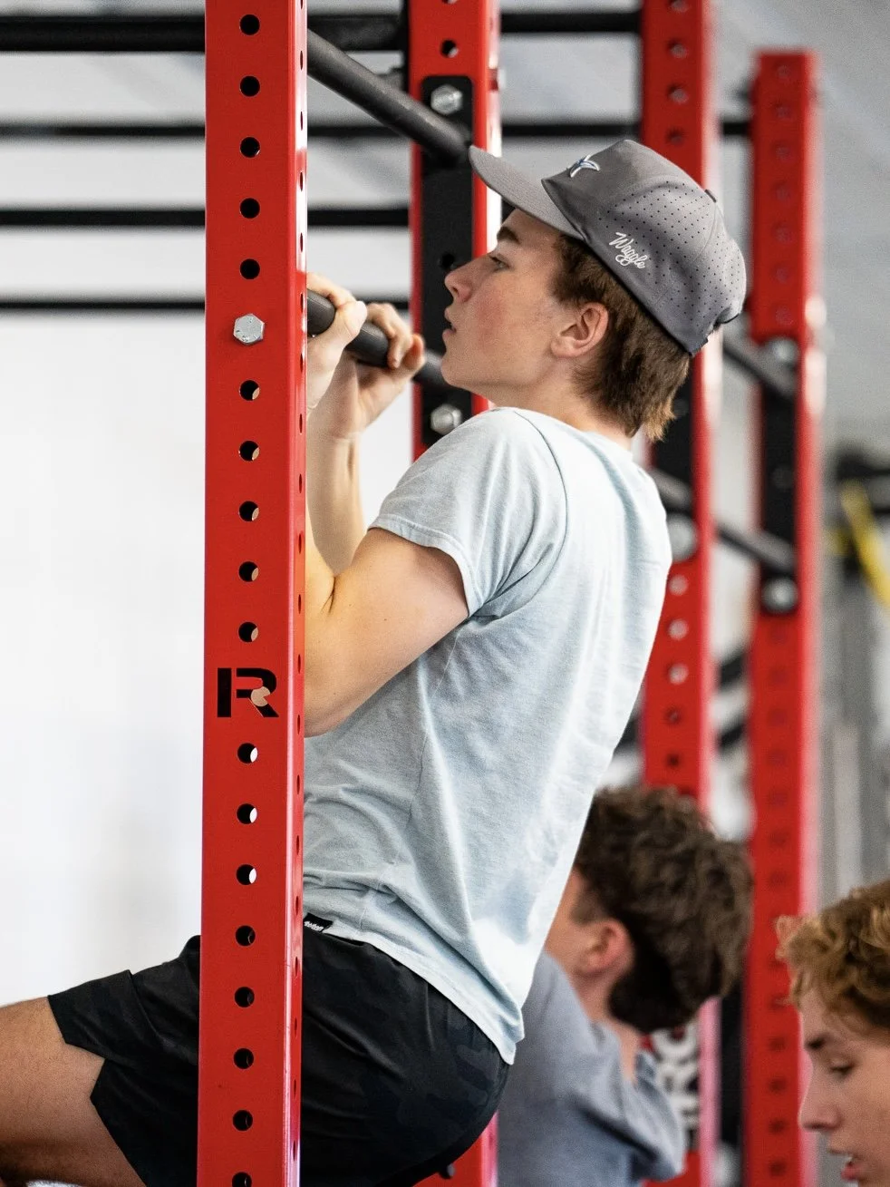 A young man wearing a grey baseball cap and light grey t-shirt doing pull-ups on a red and black gym pull-up bar in a gym with other people in the background observing or waiting.