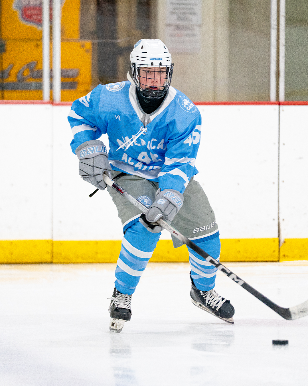 Hockey player in blue uniform with the number 45, wearing a white helmet and gray gloves, controlling the puck on the ice rink.