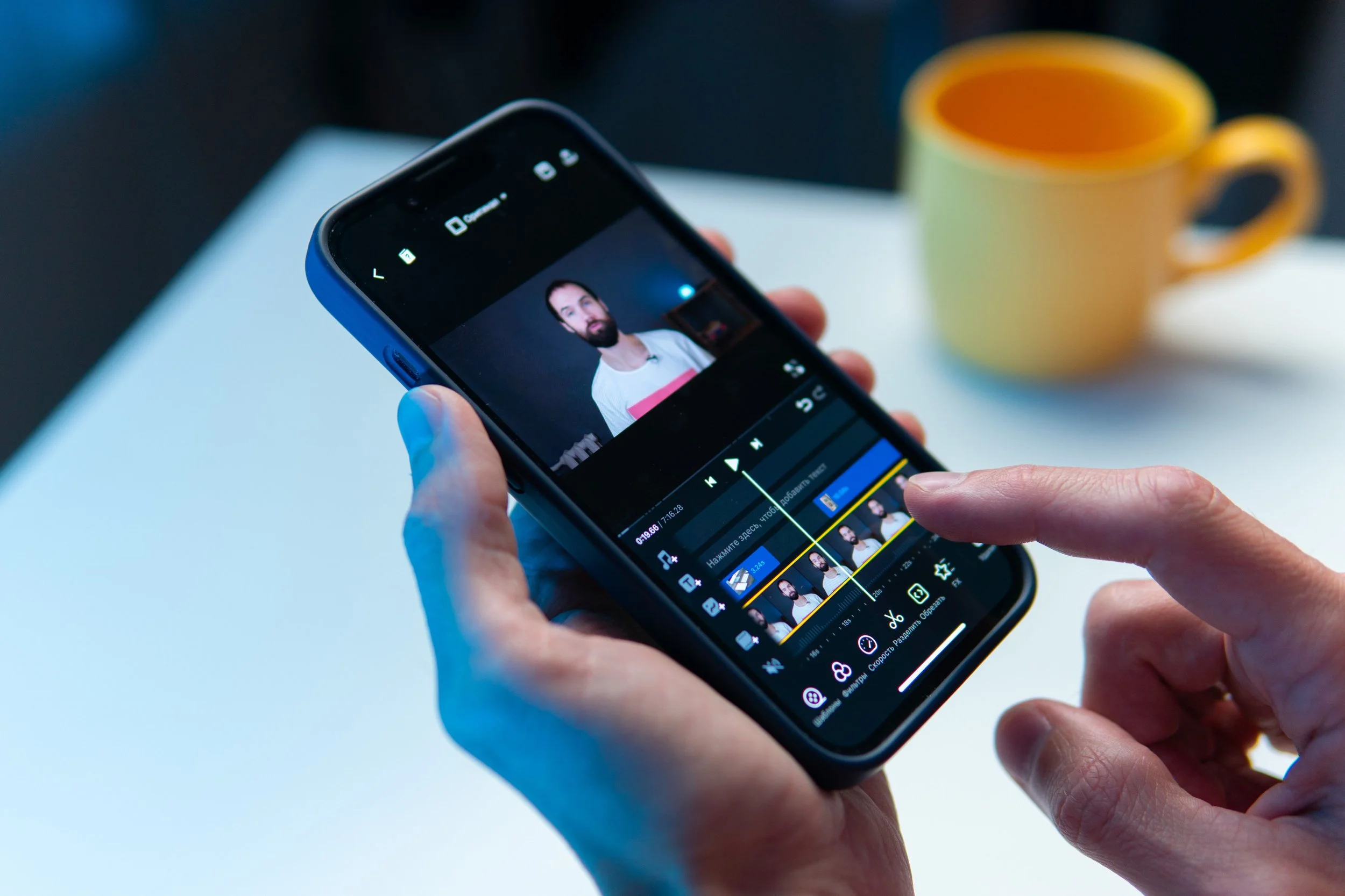 Close-up of person's hand holding a smartphone with a video editing app open, editing a photo of a man with a beard and white shirt, in a room with a yellow cup in the background.