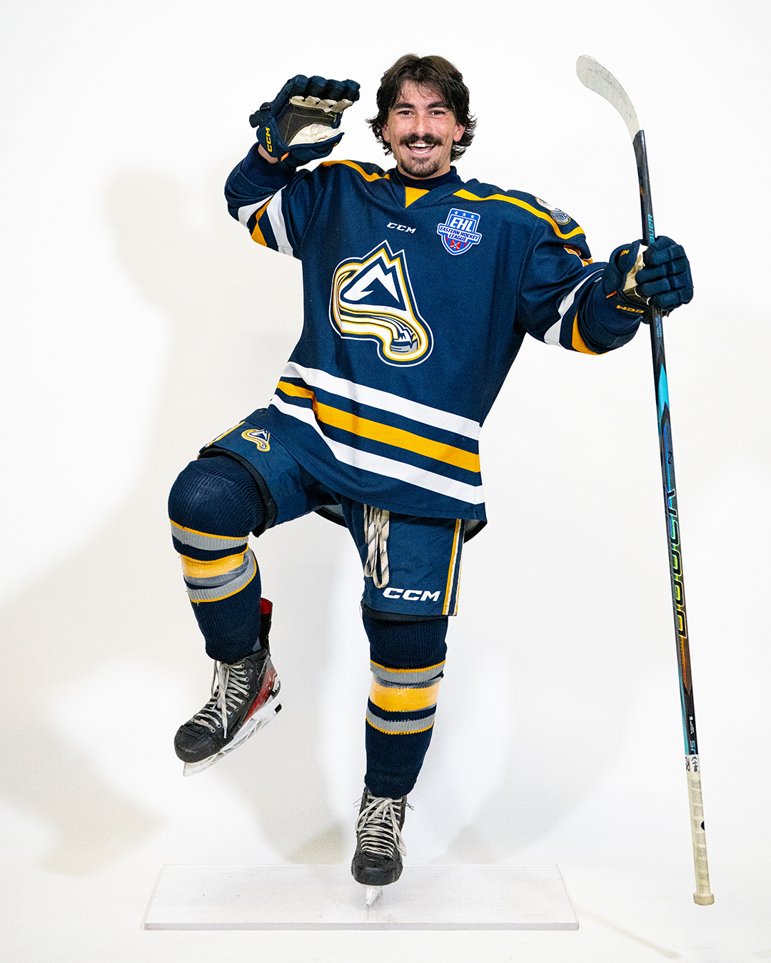 A male hockey player in a blue and yellow jersey with a large logo, holding a hockey stick, and skating on one leg against a plain white background.