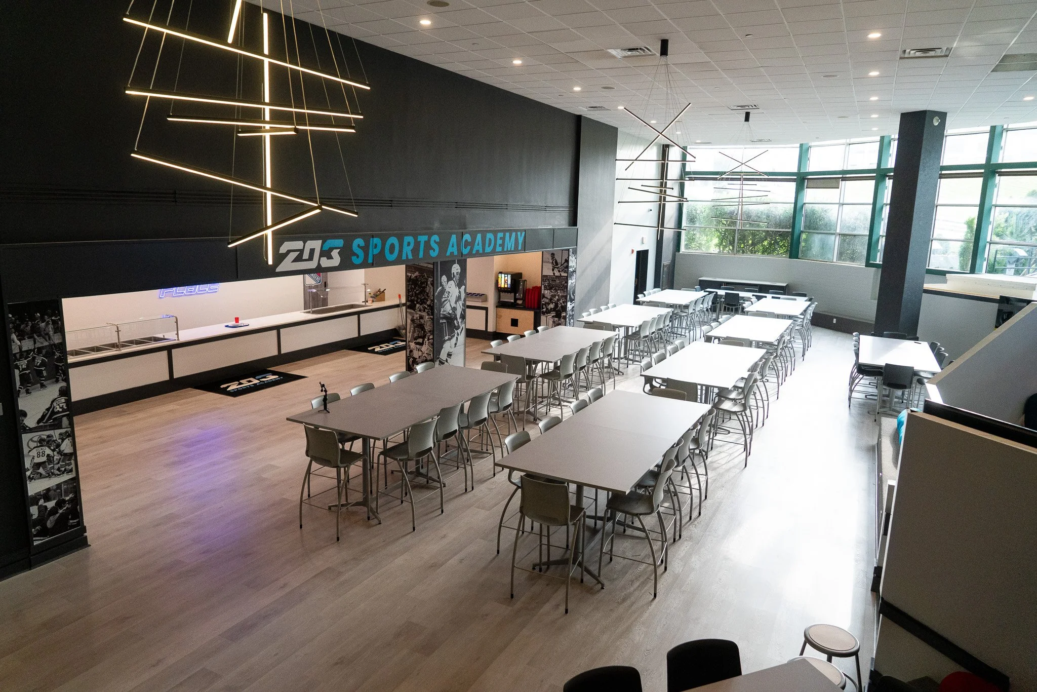 An indoor sports facility with multiple white tables and chairs arranged for dining or meetings, large windows letting in natural light, modern lighting fixtures, and a sign that reads 'Sports Academy' in the background.