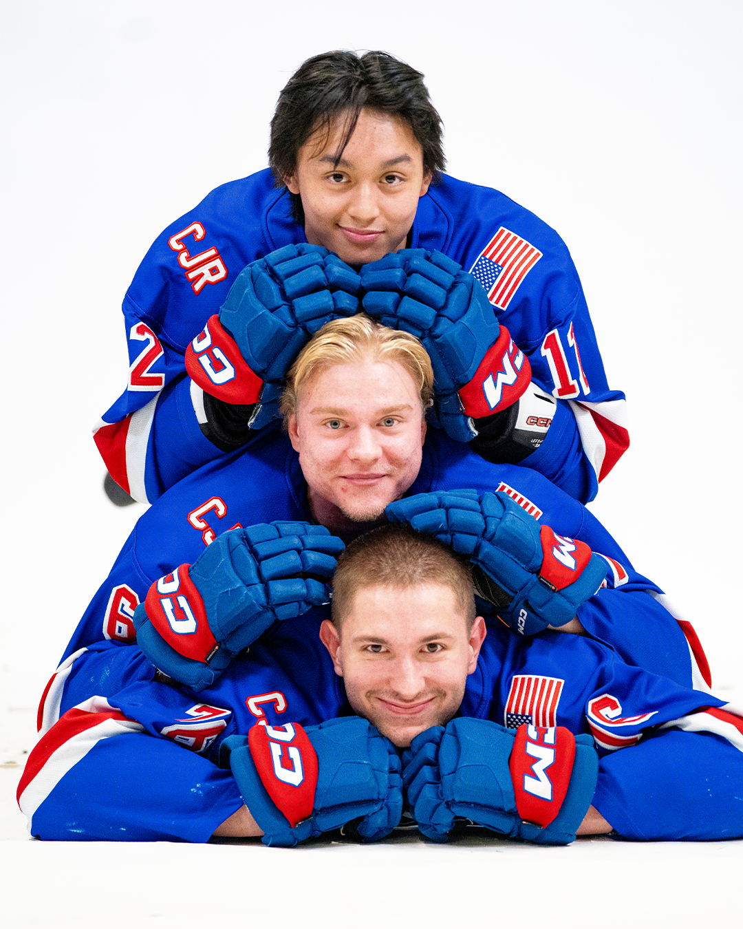Four ice hockey players in blue jerseys and gloves stacked on top of each other in a pyramid pose on the ice rink.