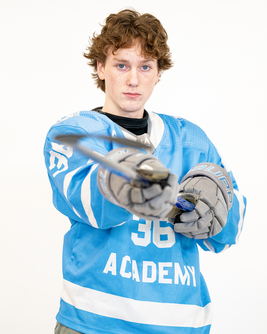 Young male hockey player in blue jersey with white lettering, pointing a hockey stick forward, wearing gray gloves, against a plain white background.