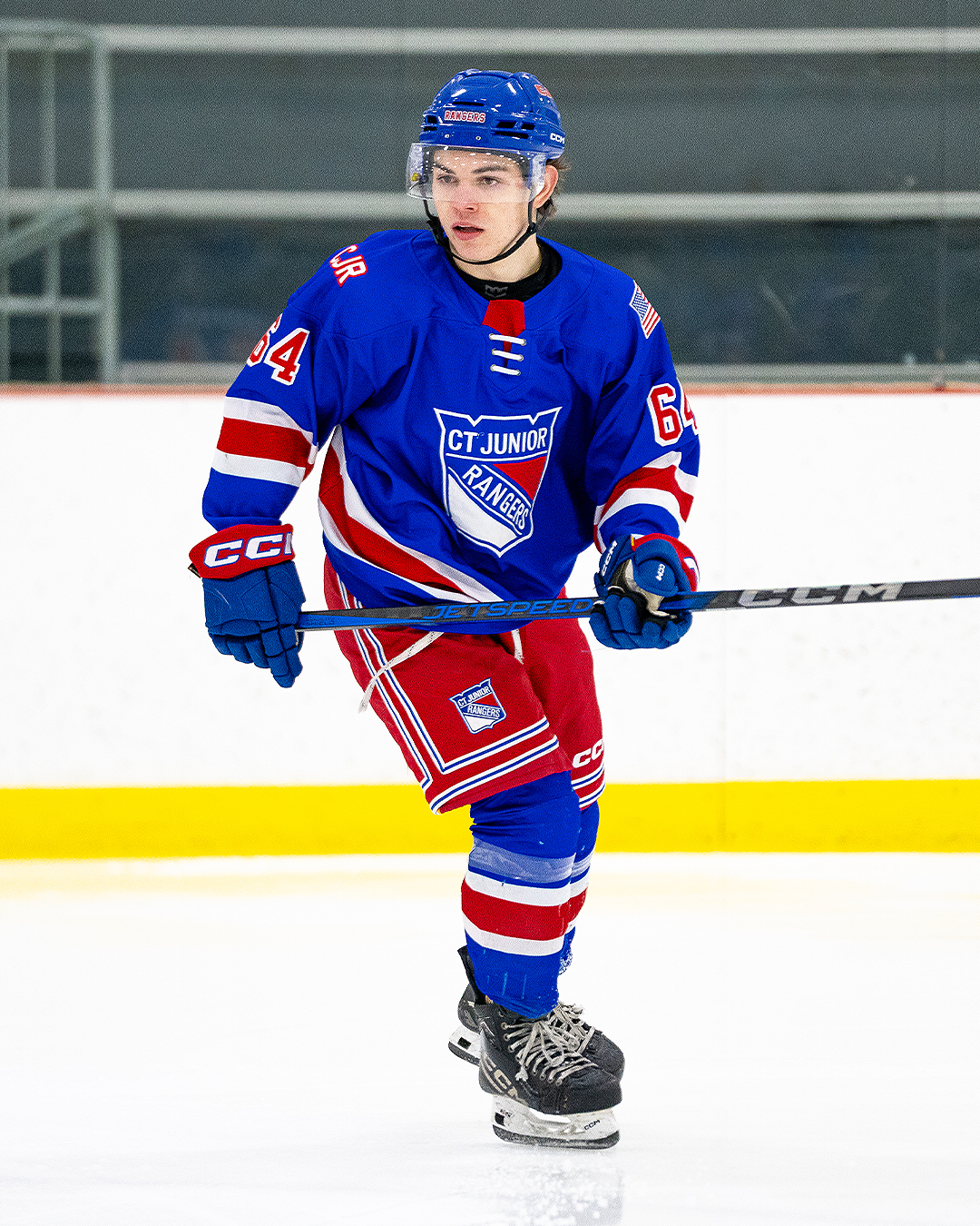Young male hockey player in blue, red, and white uniform skating on ice rink with CT Junior Rangers logo, wearing helmet and gloves, holding a hockey stick.