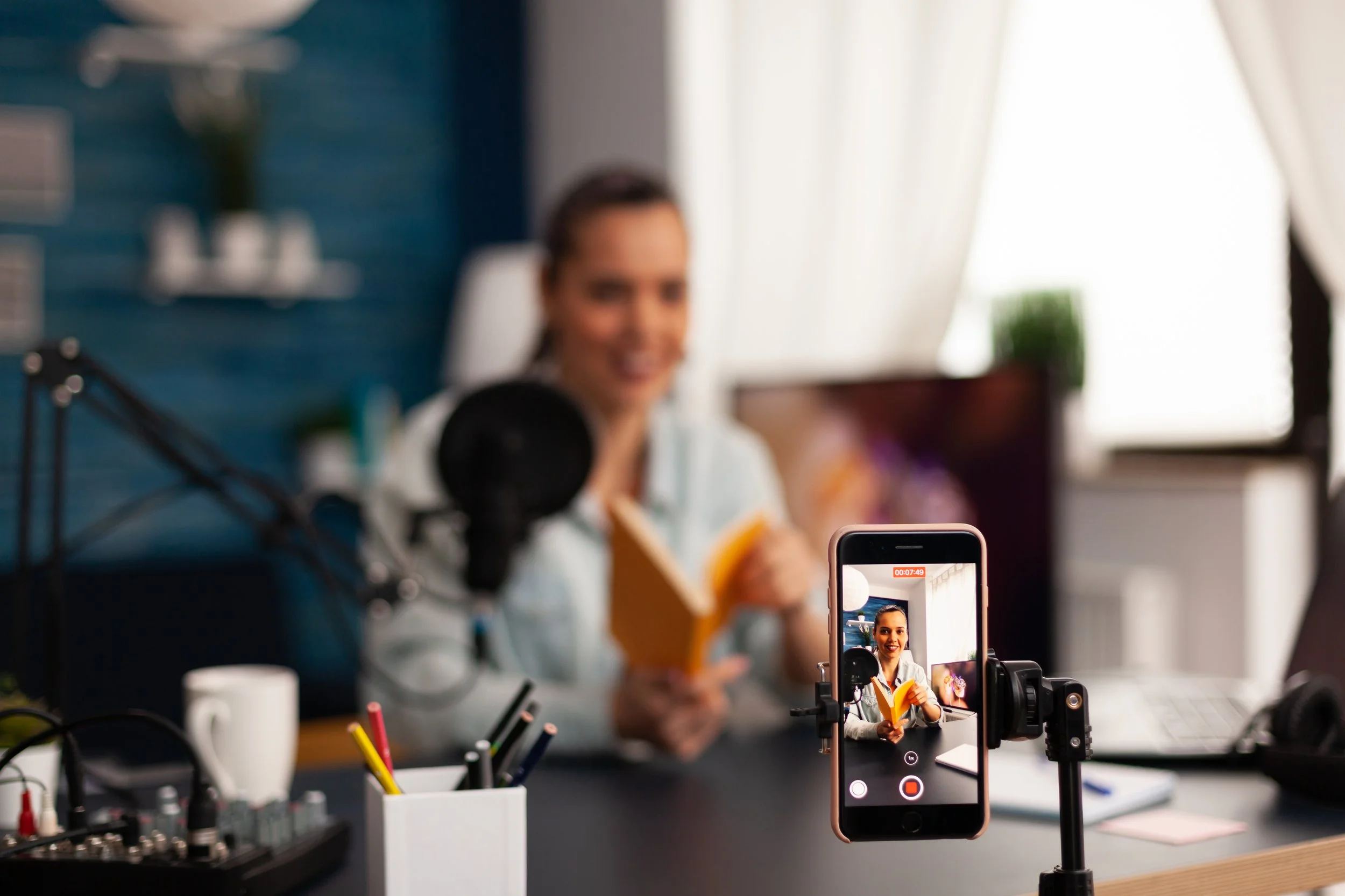 A woman recording a video with a smartphone setup on a tripod, with a microphone and audio equipment on a desk in front of her. She is smiling and appears to be in a well-lit room with a window and curtains in the background.