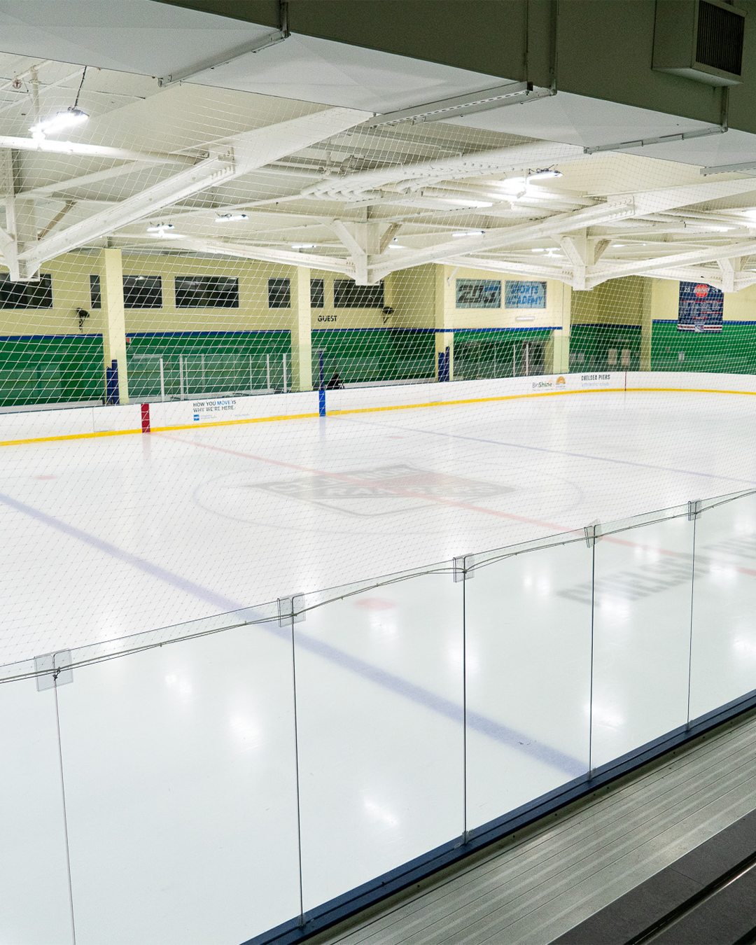 An empty indoor ice hockey rink with a glass barrier in the foreground and green and white walls in the background.