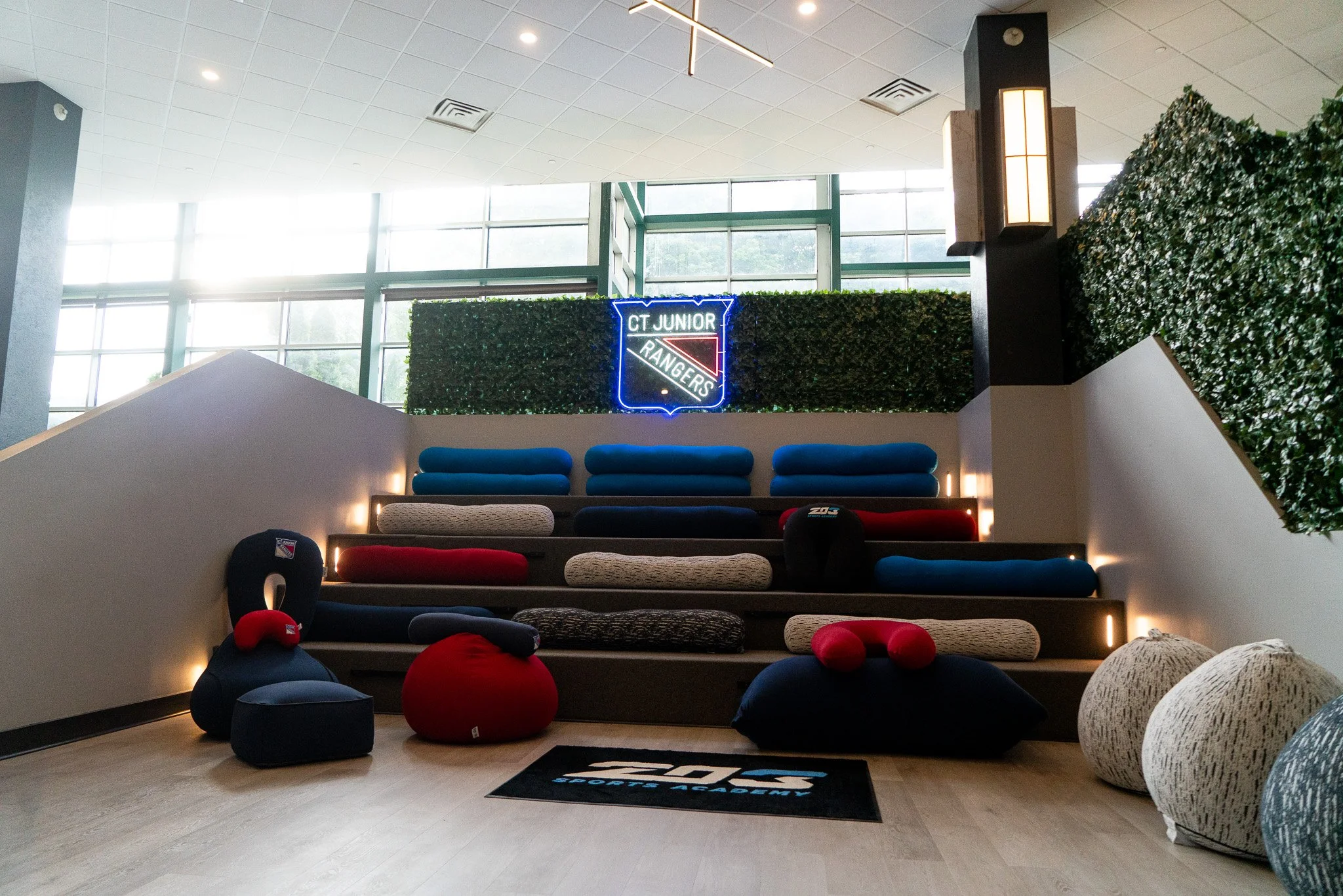 Indoor sports or fitness lounge with tiered seating filled with bean bag chairs and cushions, large windows, a neon sign reading 'CT Junior Rangers', and decorative greenery on the walls.