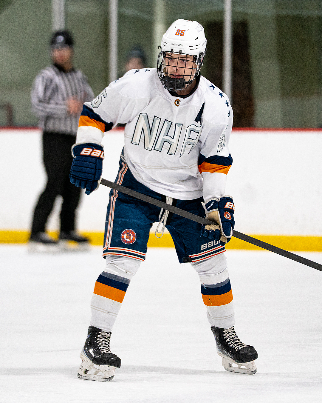 Ice hockey player in white jersey with NHFC logo, wearing a helmet, gloves, and skates, holding a hockey stick, on an ice rink with a referee in background.