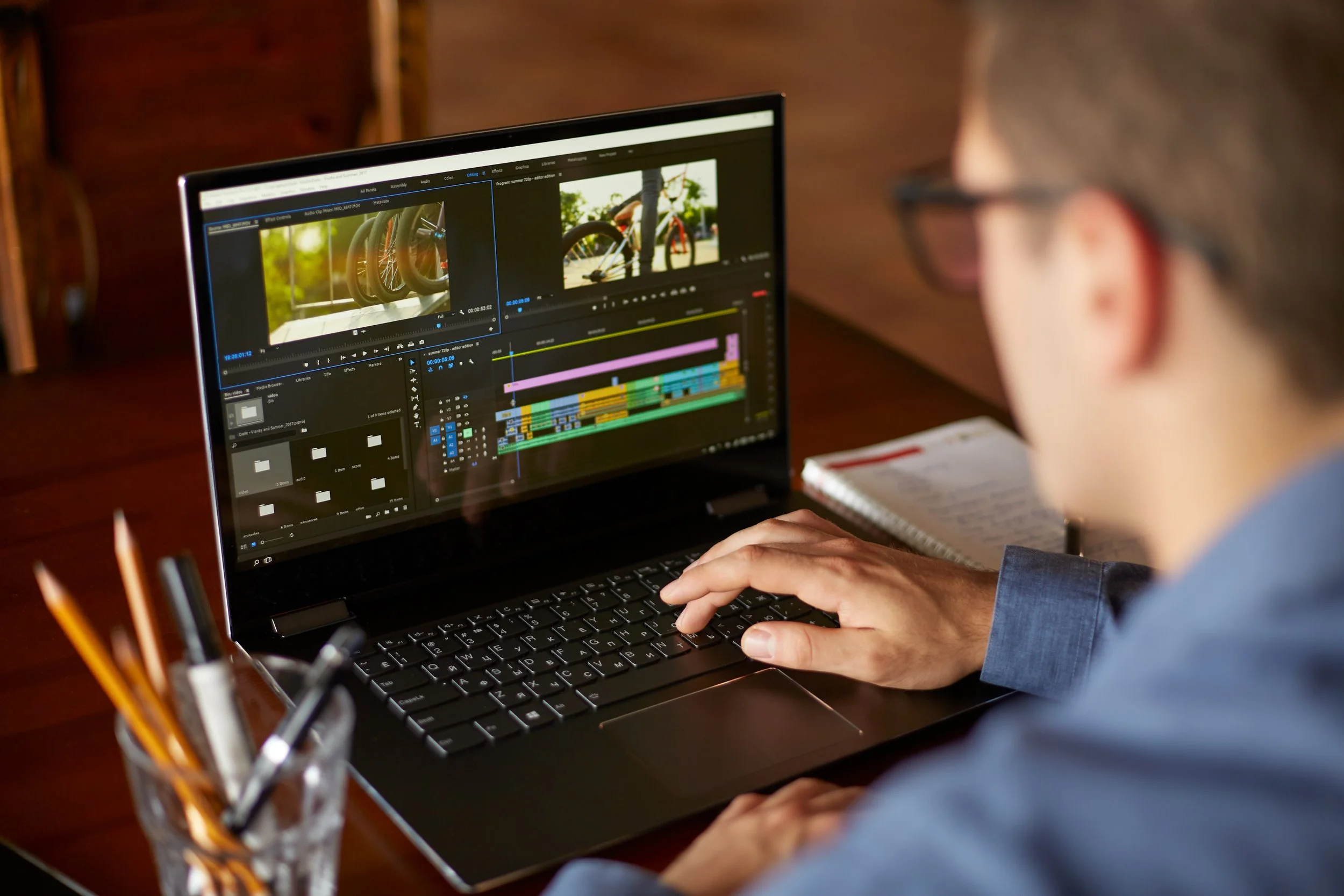A person working on a laptop editing a video with a timeline and preview windows open, sitting at a desk with a notebook and a jar of pencils.