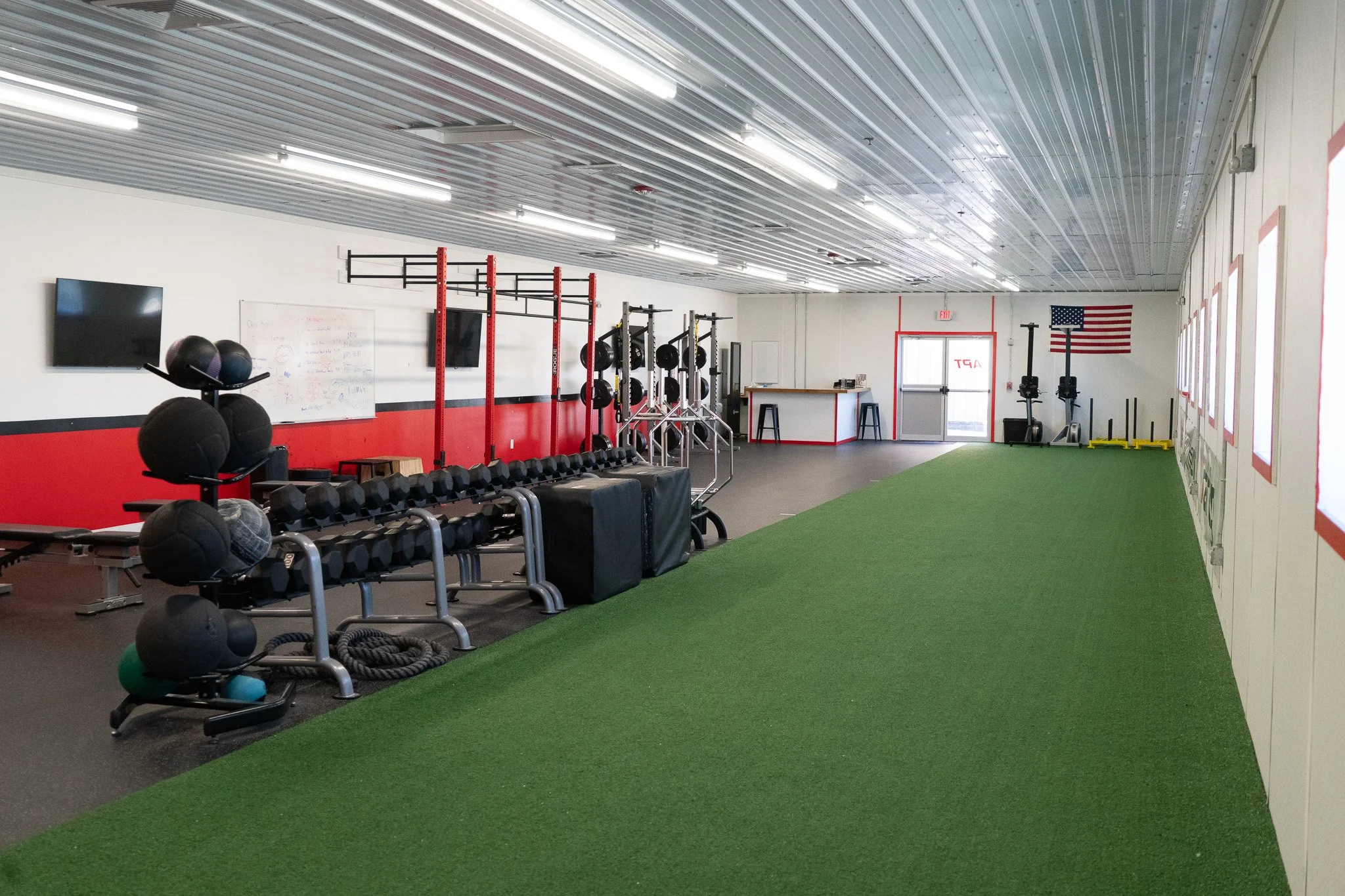 Interior of a gym with workout equipment including medicine balls, dumbbells, squat racks, rowing machines, and a green turf area, with an American flag on the wall.