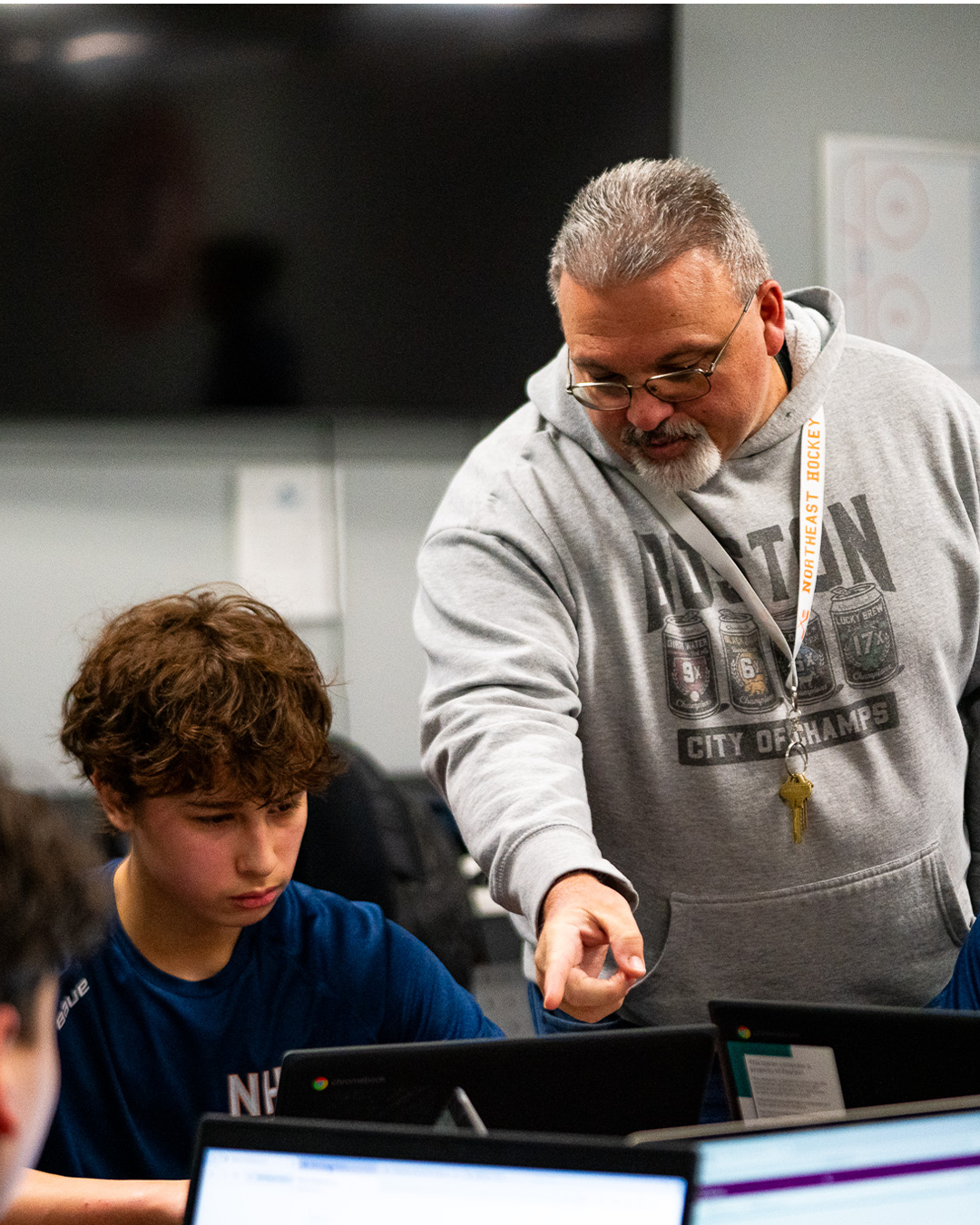 An older man with glasses and gray hair helps a young boy with curly hair using a laptop in a classroom or computer lab.