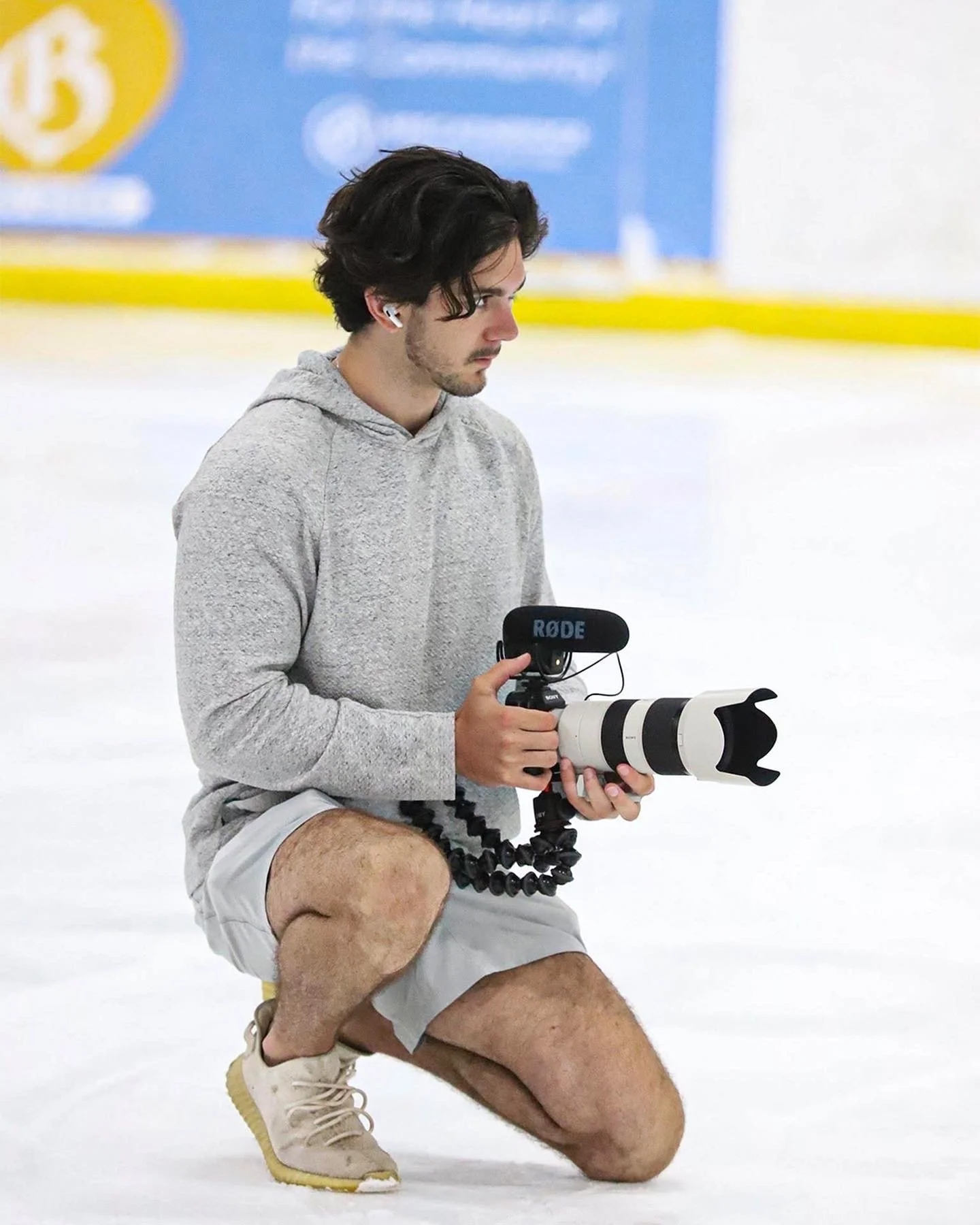 Young man kneeling on one knee on an ice rink, holding a professional camera with a large lens and microphone, wearing a gray hoodie, shorts, and sneakers.