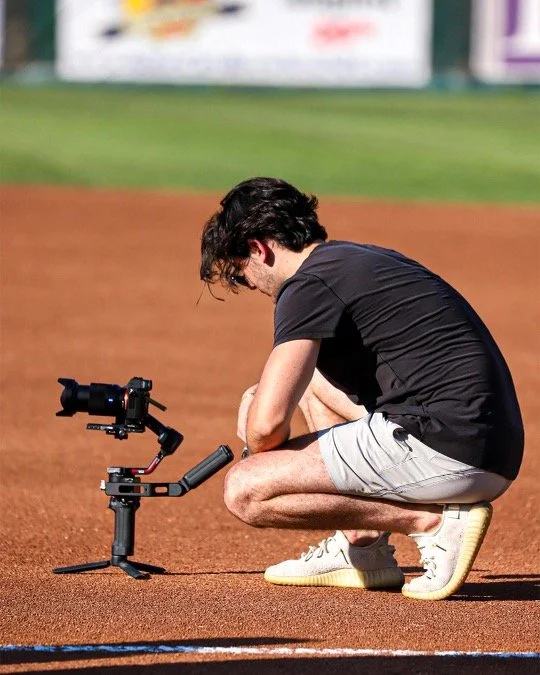A man crouching on a baseball field with a camera mounted on a tripod in front of him.