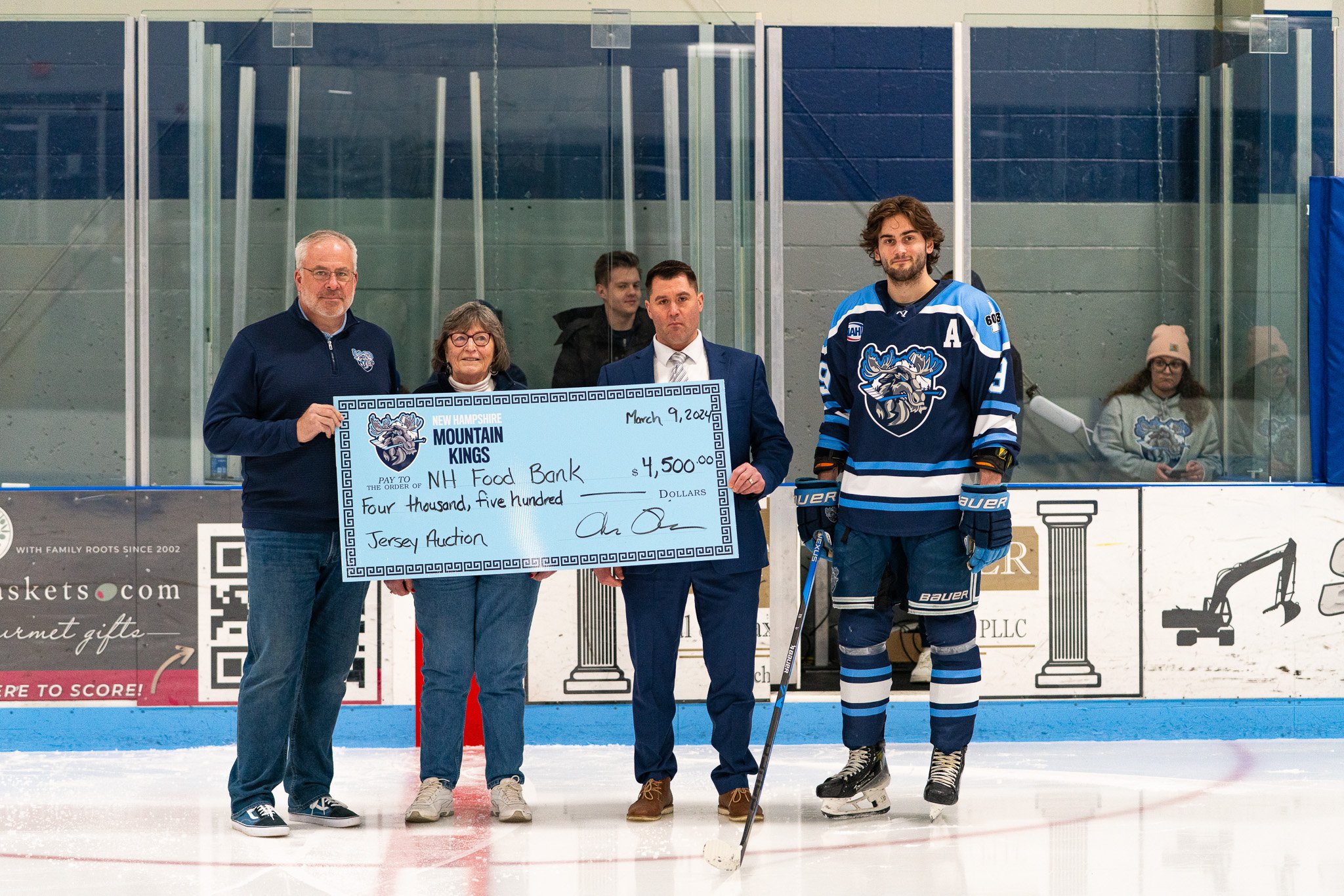 A group of five people on an ice hockey rink holding a large check, with a hockey player in uniform standing on the right. The check is addressed to the NH Food Bank for $4,500, dated March 9, 2024. The group appears to be participating in a charity event.