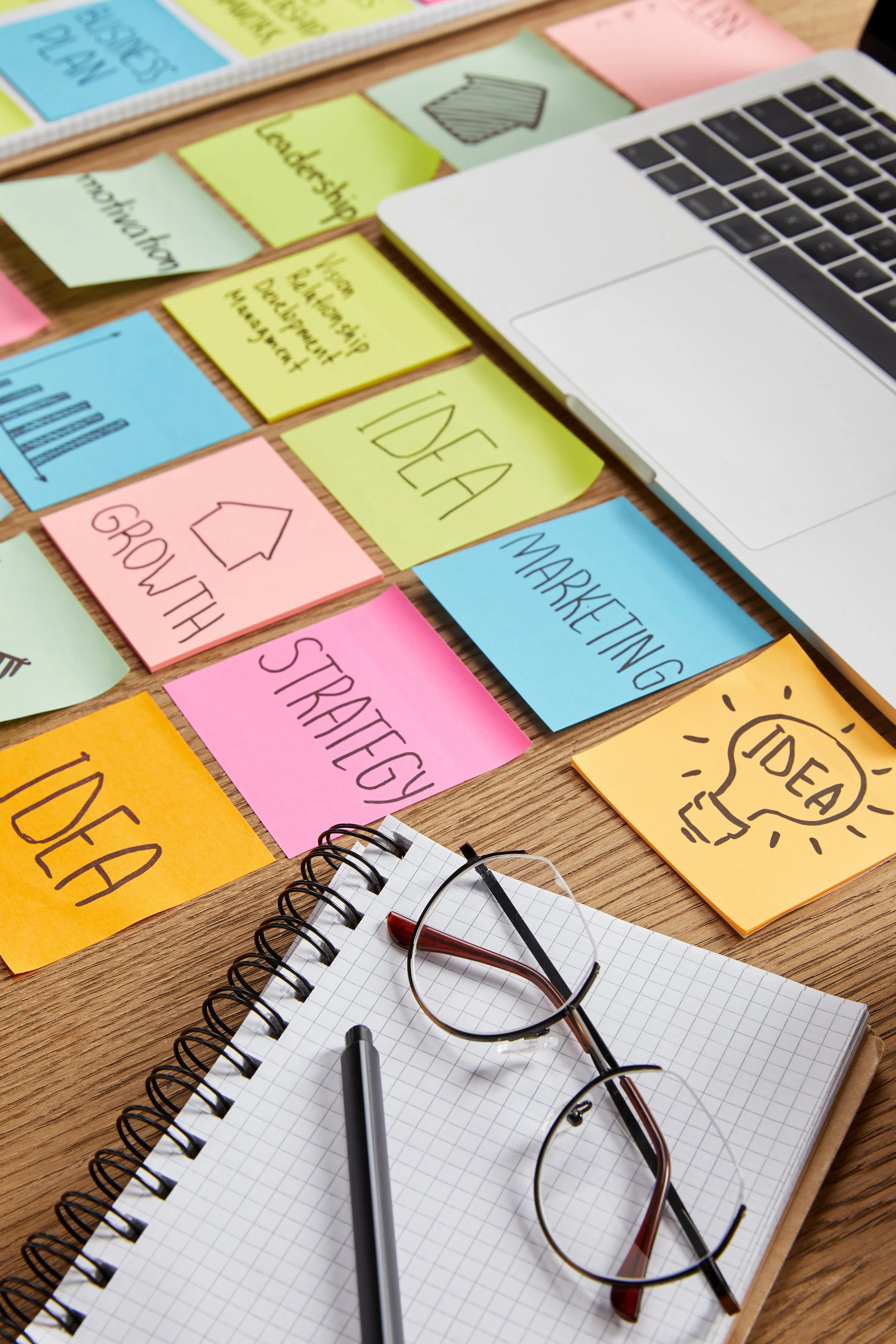 Office desk with colorful sticky notes containing business-related words like 'IDEA,' 'MARKETING,' and 'GROWTH,' a spiral notebook with a pen, and eyeglasses, along with part of a laptop.