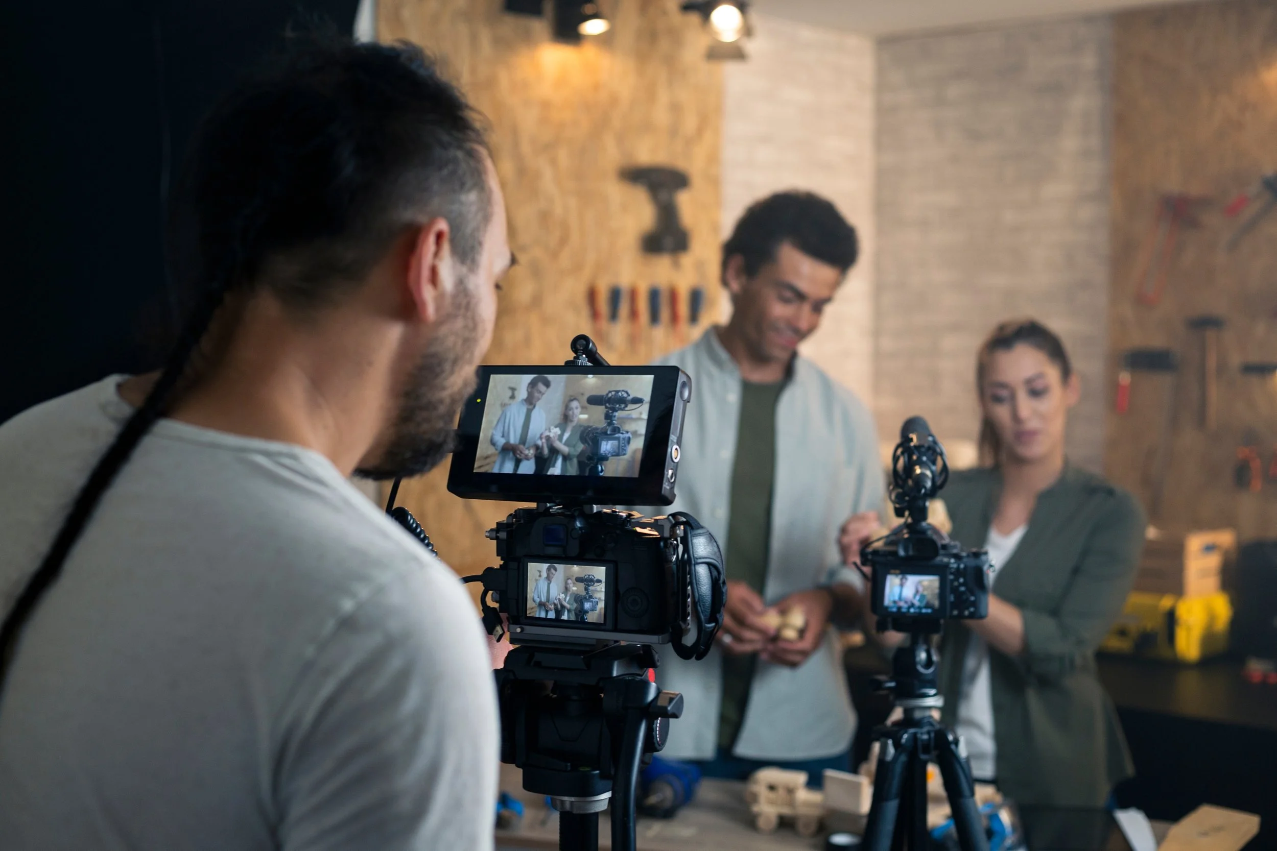 A man operating a camera with a monitor screen showing two people speaking in front of a brick wall with tools and equipment in the background.